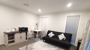 A compact living space with a kitchenette, a black sofa with floral cushions, a patterned rug, and a window with blinds.