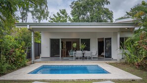 Una casa moderna con piscina, un patio con mesa y sillas, y un jardín con diversas plantas.
