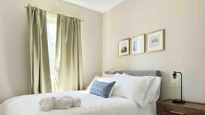 A neatly made bed with white linens and a blue patterned pillow, a wooden nightstand with a lamp, and a window with beige curtains. - Spring Street Suites A / Stylish 3BD/1BA Apt (Evansville)