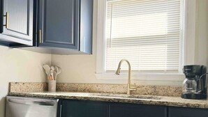A kitchen with dark cabinets, a stainless steel dishwasher, a granite countertop, a window with blinds, and a coffee maker. - Spring Street Suites A / Stylish 3BD/1BA Apt (Evansville)