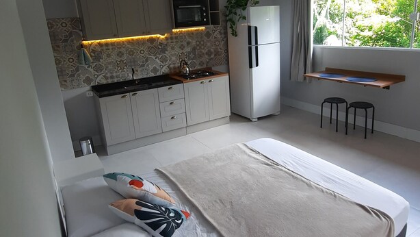 A modern bedroom with a large bed, a small kitchenette, and a window with a view of greenery. - Studio Flor do Mato (Itapema)