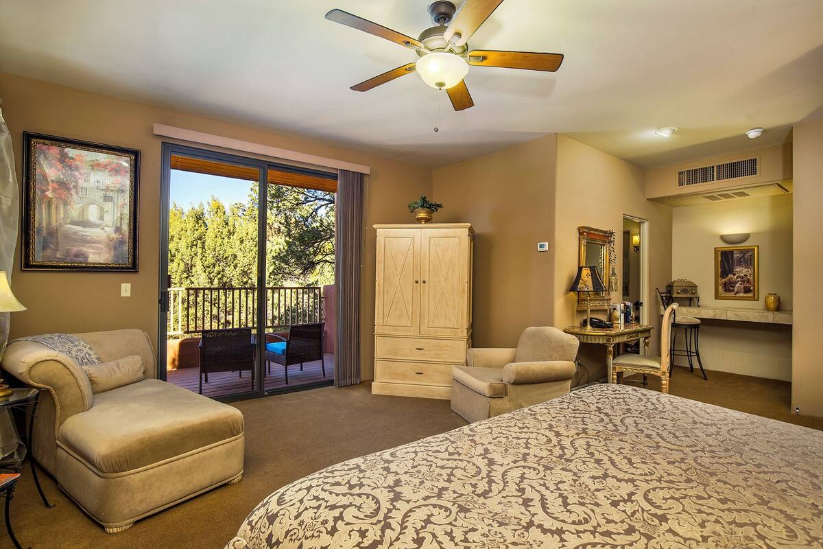 A bedroom with a large bed, a ceiling fan, a sliding glass door leading to a balcony, a wardrobe, a desk with a chair, and a mirror.