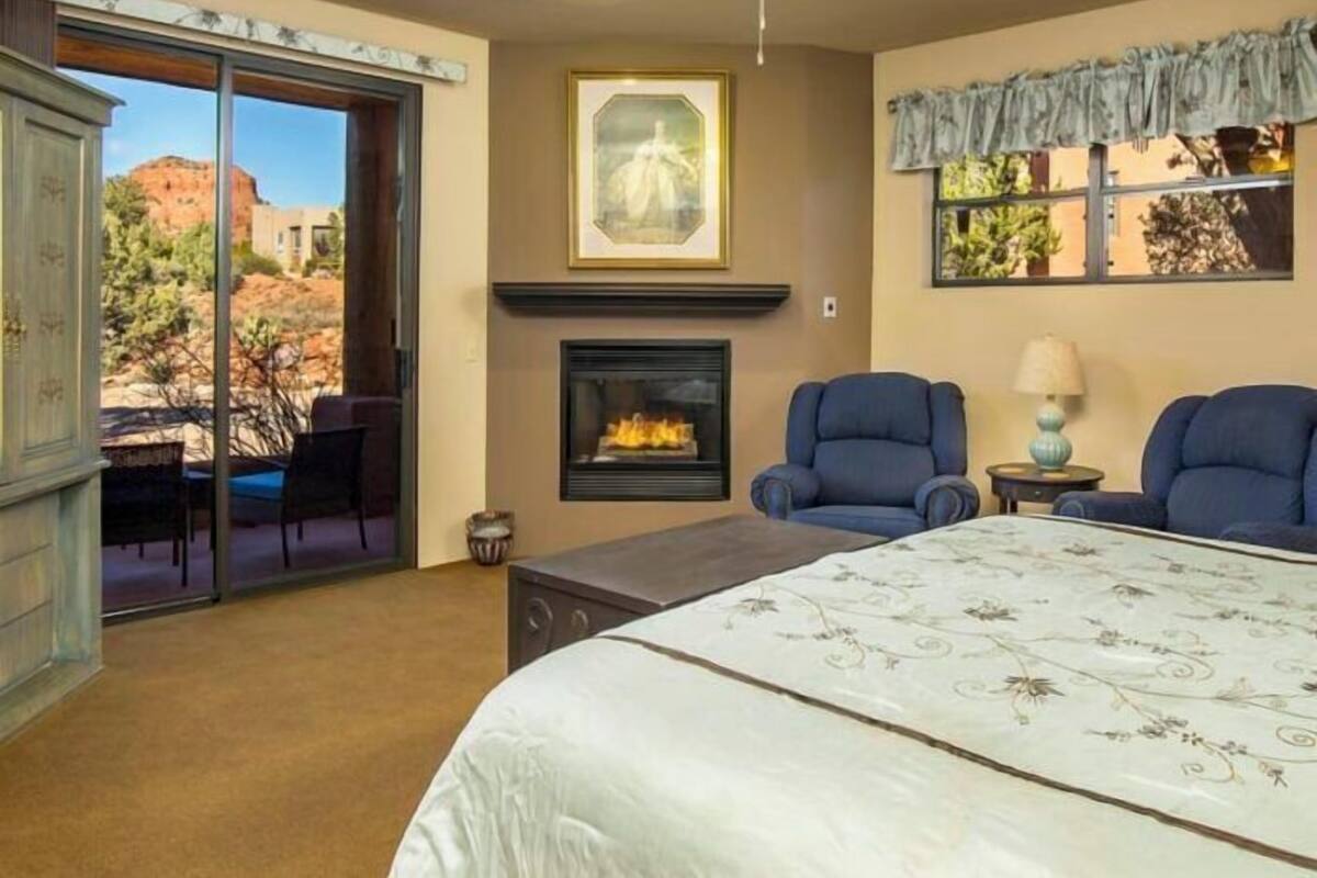 A bedroom with a bed, two armchairs, a fireplace, and a large window with a view of a rocky landscape.