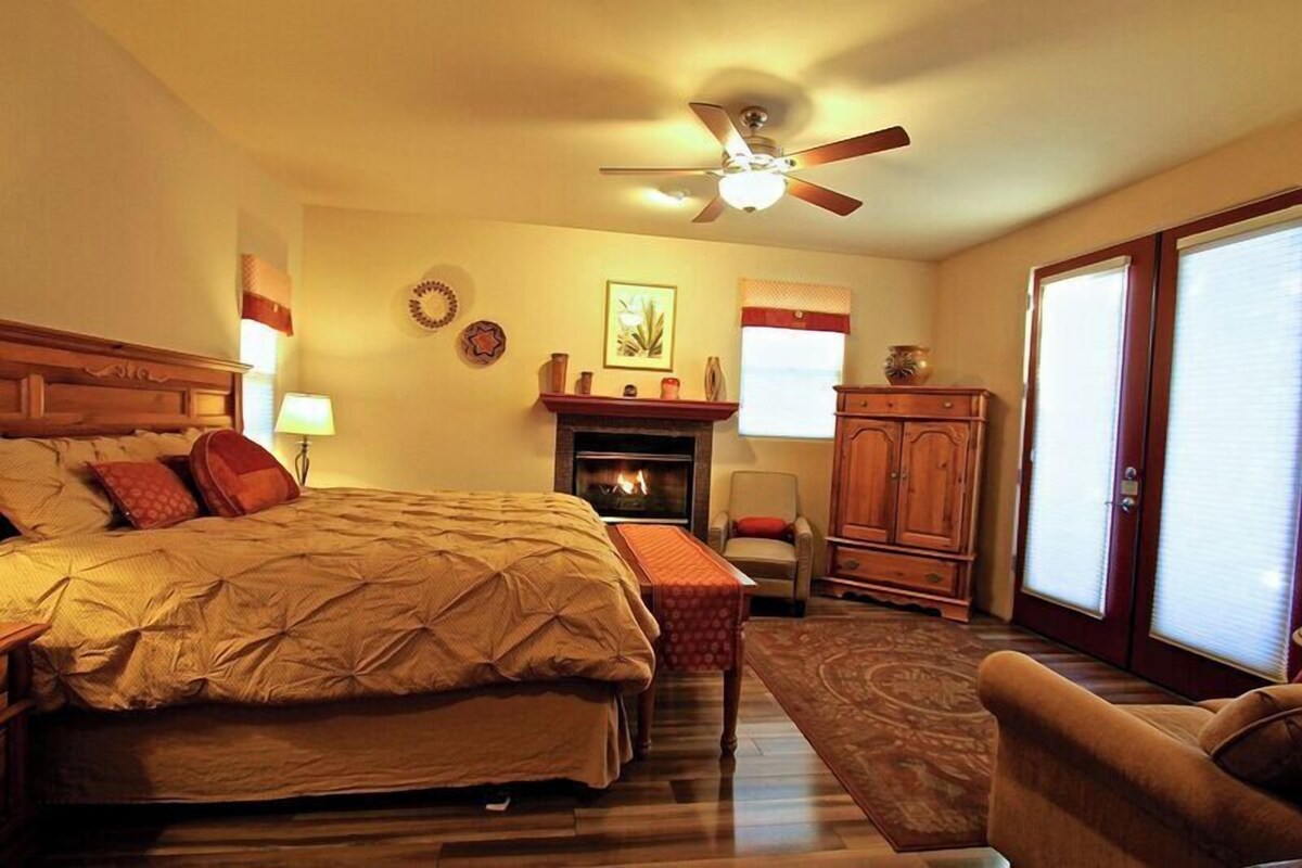 A bedroom with a large bed, a fireplace, a ceiling fan, and a view of the outside through a window.