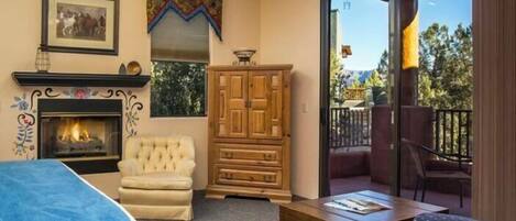 A cozy room with a fireplace, a cushioned chair, a wooden cabinet, and a balcony with a view of trees and mountains.