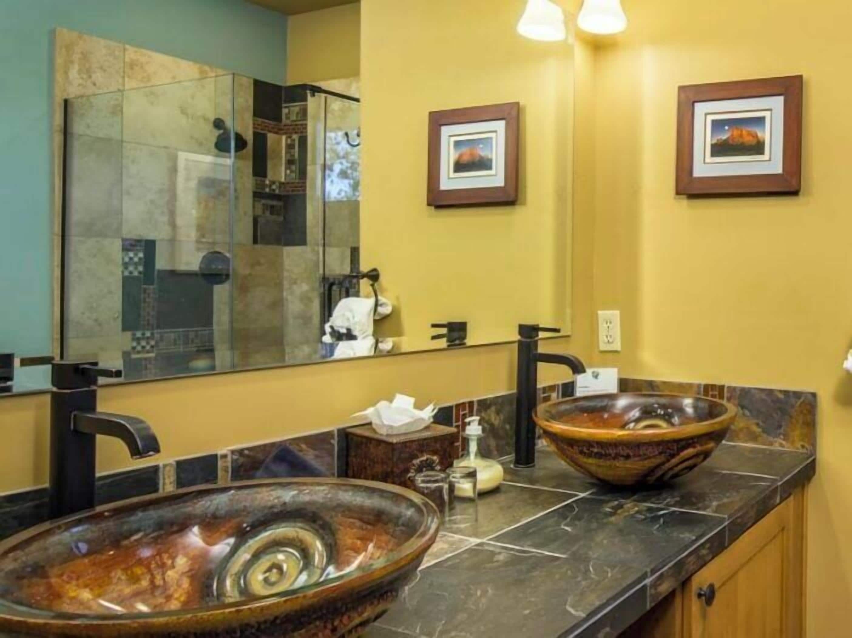 A bathroom with two large, oval-shaped ceramic sinks, a black faucet, a mirror with a tiled backsplash, and two framed pictures on the wall.