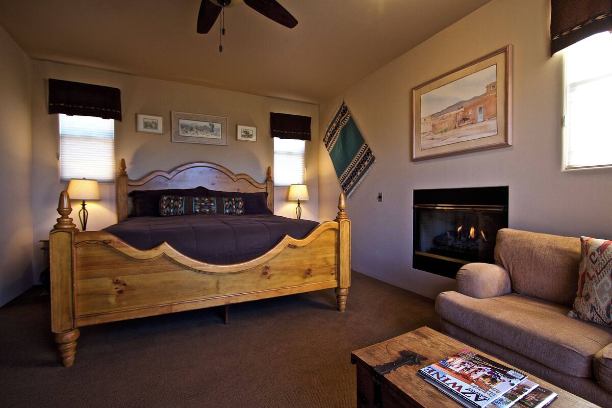 A bedroom with a wooden bed, two bedside lamps, a sofa, a fireplace, and framed pictures on the walls.