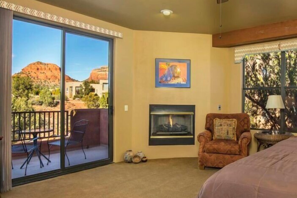 A room with a fireplace, a chair, a bed, and a balcony with a view of a desert landscape.