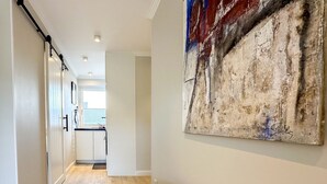 A hallway with a kitchen area visible through an open door, a large abstract painting on the wall, and a wooden floor. - Flow - Haus Alice - Flow (Sylt)