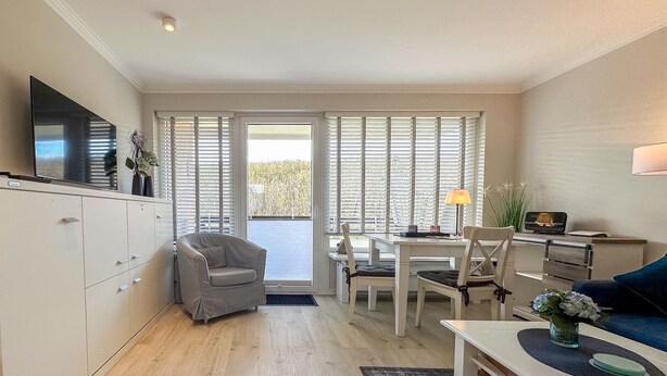 A compact living space with a dining area, a sofa, a TV, and a window with blinds. - Flow - Haus Alice - Flow (Sylt)