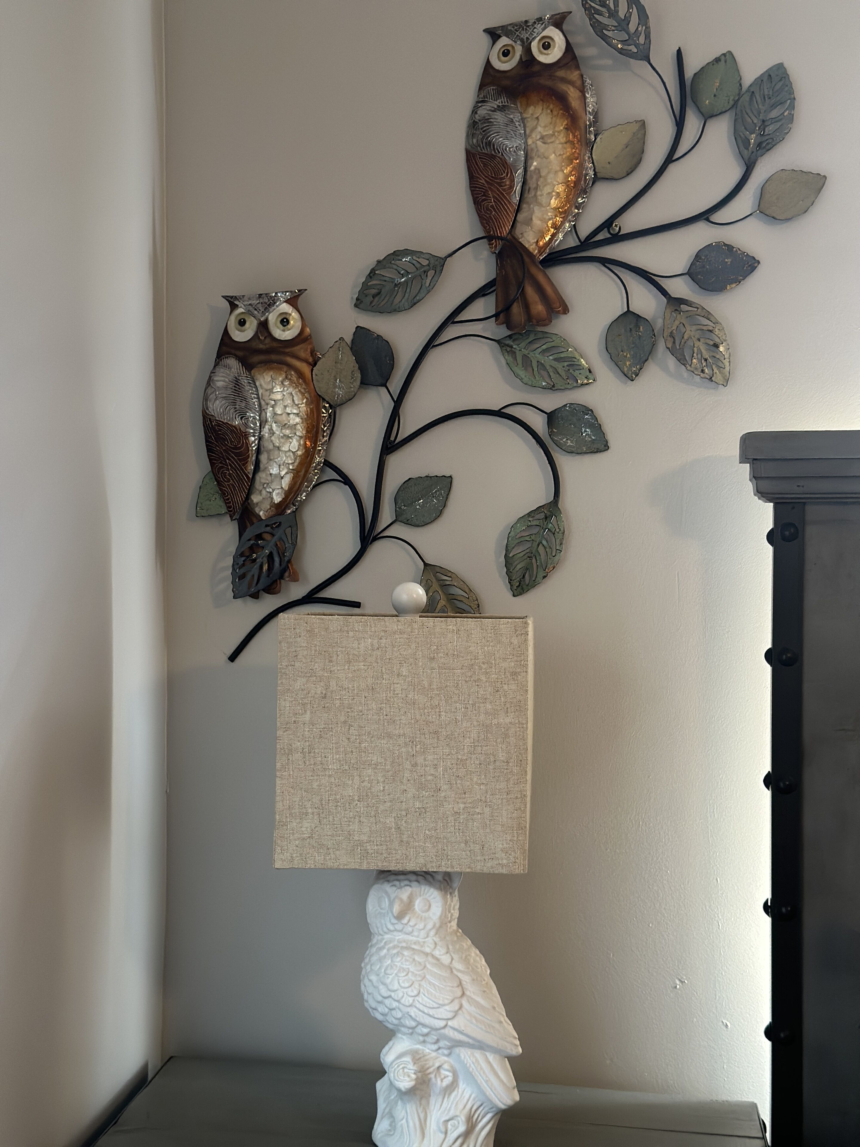 A wall-mounted metal art piece of two owls on a branch, a textured lamp, and a white sculptural owl base.