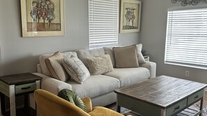 A living room with a sofa, armchair, coffee table, and side table. There are two windows with blinds, a framed artwork on the wall, and a small table with a lamp.