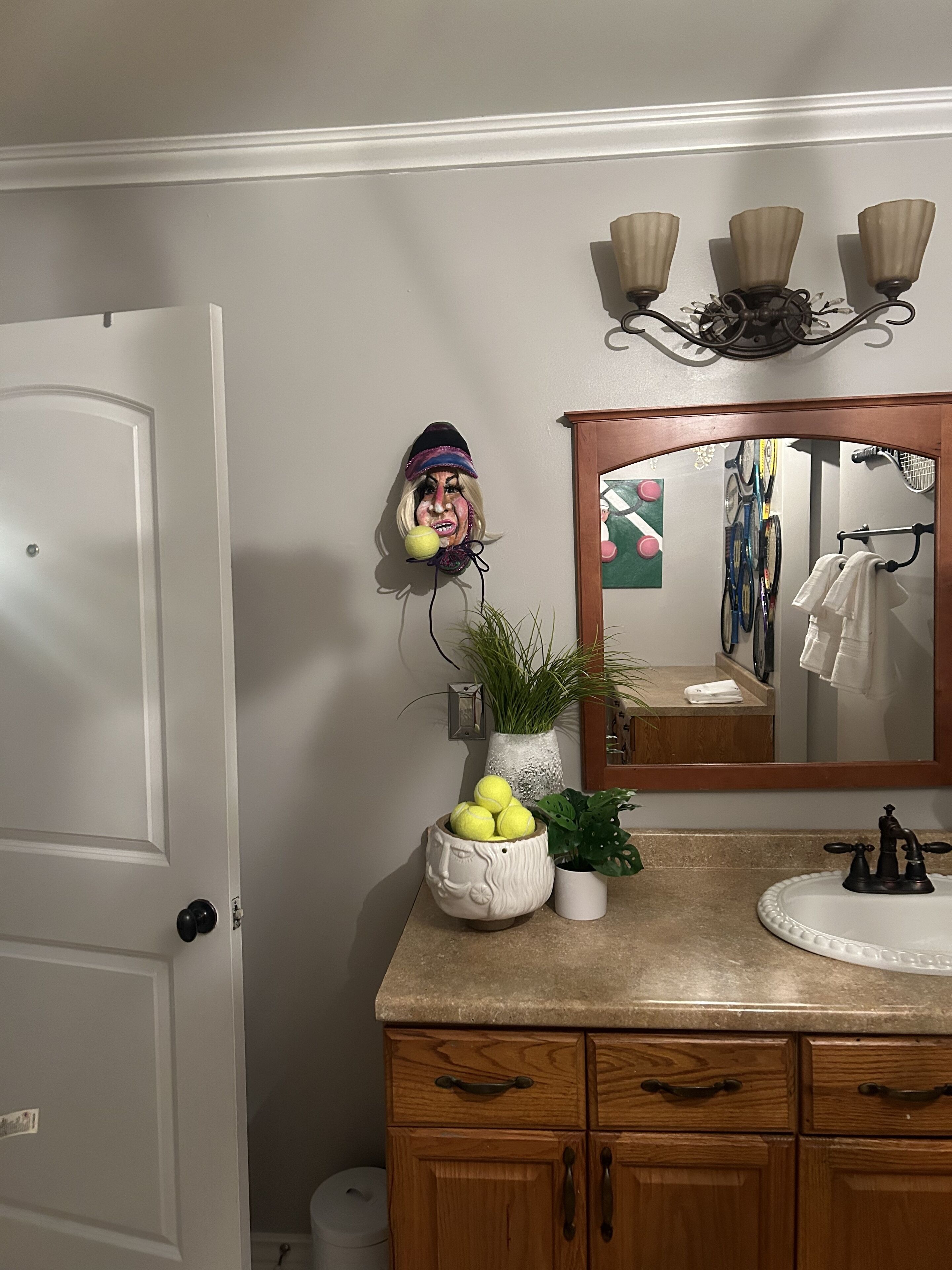 Baño con tocador de madera, espejo y lavabo. Hay elementos decorativos como una planta en maceta y un jarrón con flores amarillas.