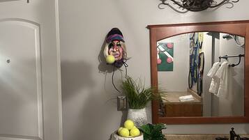 Baño con tocador de madera, espejo y lavabo. Hay elementos decorativos como una planta en maceta y un jarrón con flores amarillas.