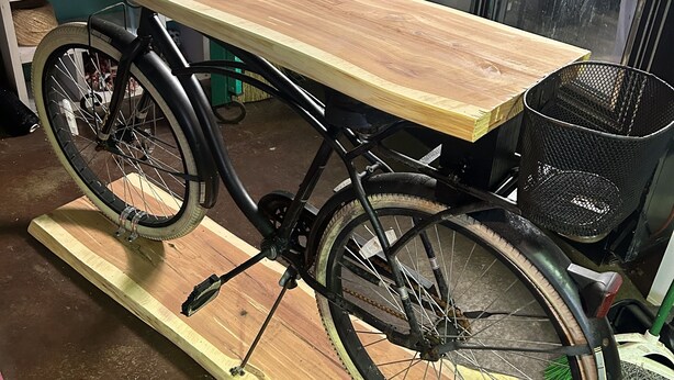 A bicycle with a wooden table attached to its frame, placed on a wooden platform in a room with green walls and various items in the background.