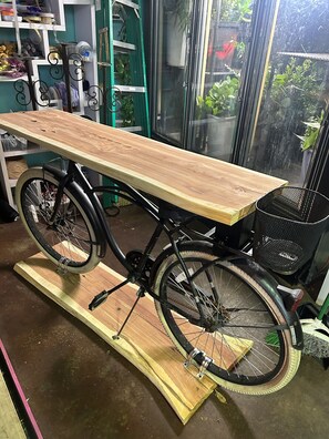 Una bicicleta con una mesa de madera unida a su estructura, sobre una plataforma de madera en una habitación con paredes verdes y varios objetos al fondo.