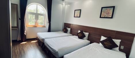 A hotel room with two beds, a large window with curtains, wooden flooring, and framed artwork on the wall.