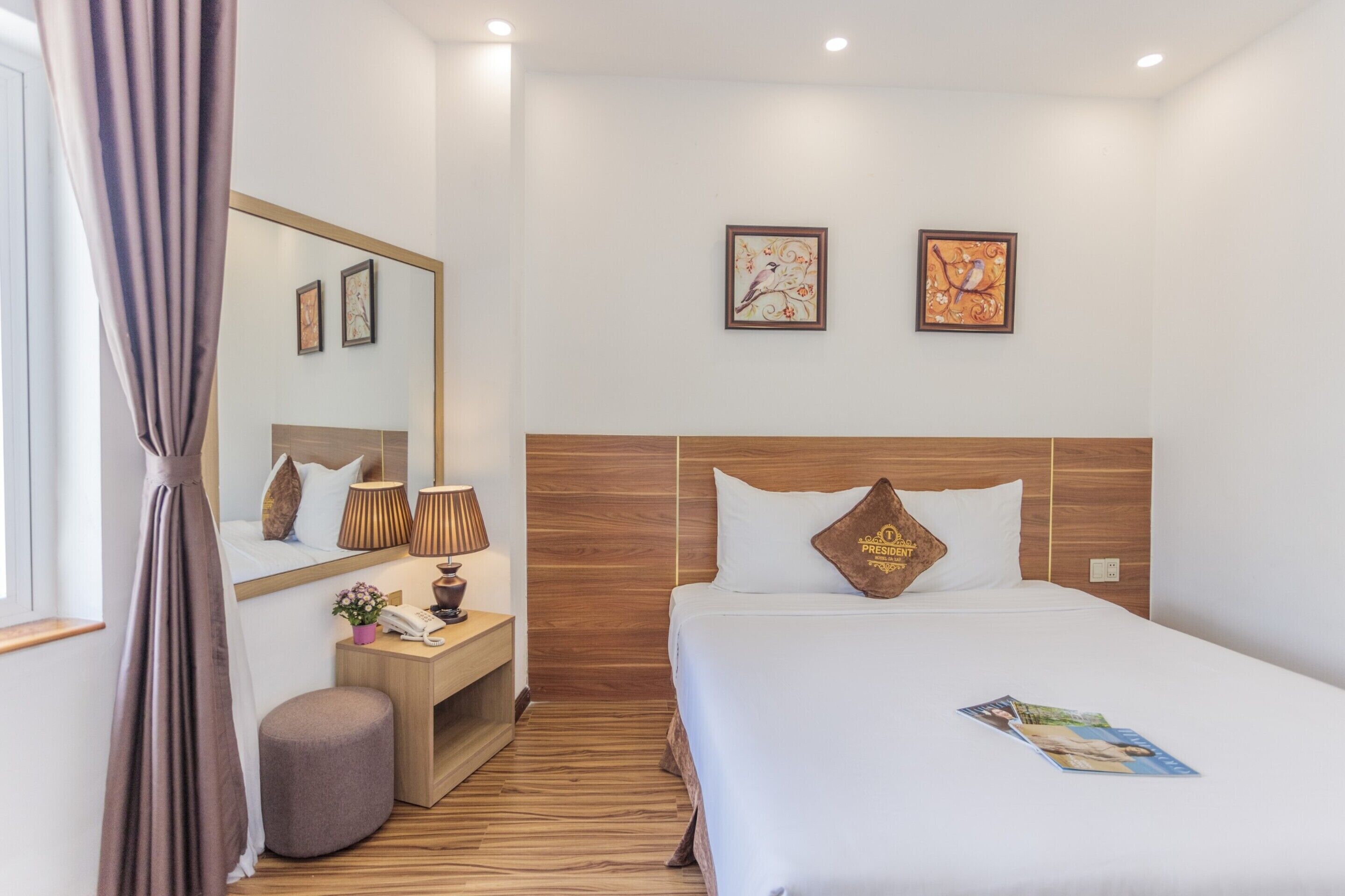 Een hotelkamer met een houten hoofdeinde, een grote spiegel, een nachtkastje met een lamp, een klein rond poefje en twee ingelijste kunstwerken aan de muur.