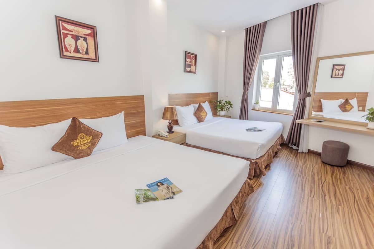 A hotel room with two beds, a wooden floor, a desk with a mirror, and a window with curtains.