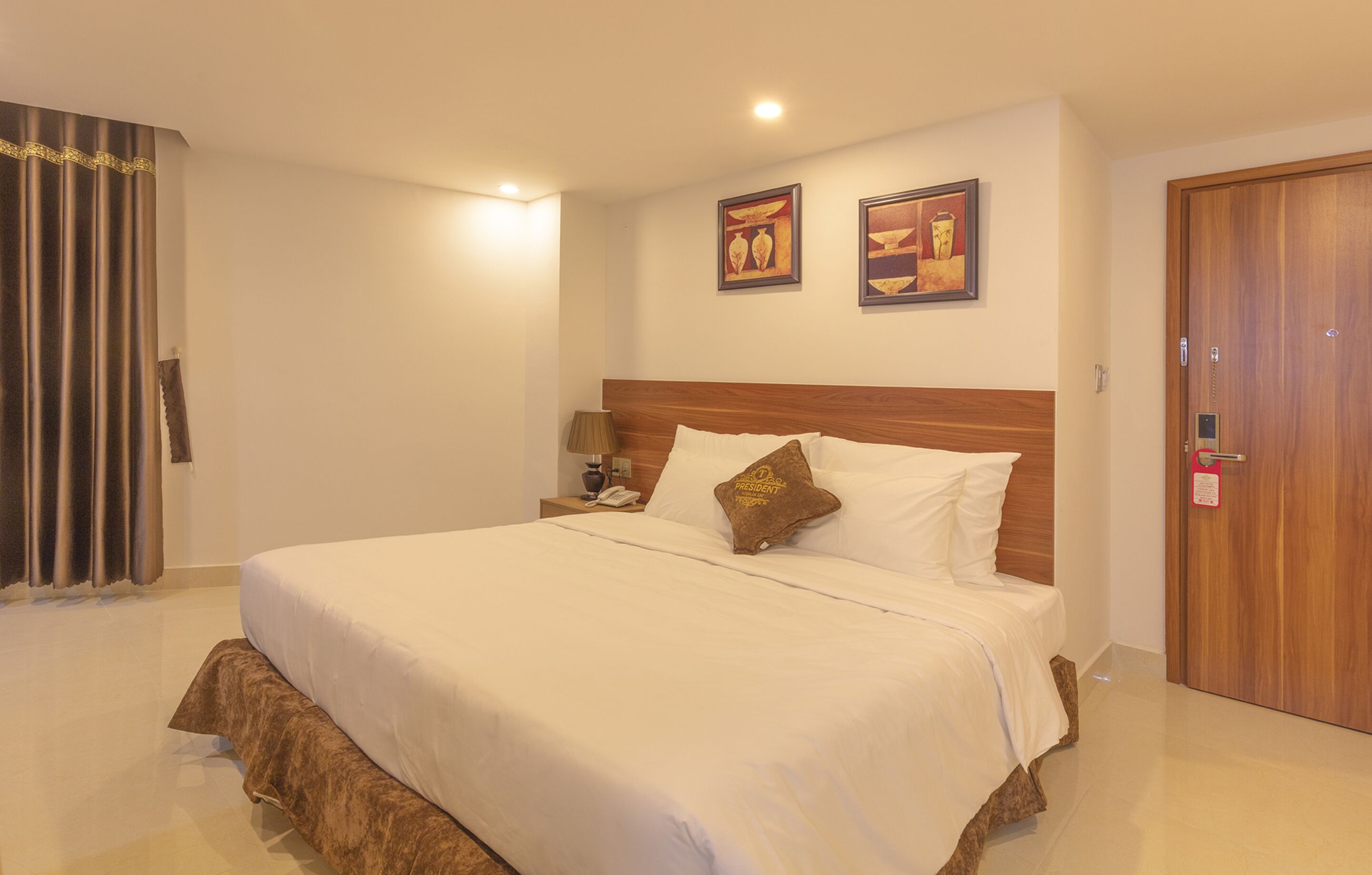 A hotel room with a large bed, a wooden headboard, two framed pictures on the wall, a bedside table with a lamp, and a door with a lock.