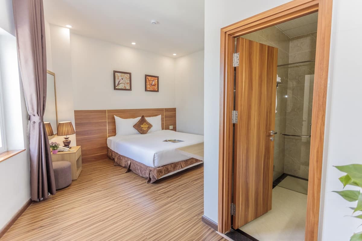 A hotel room with a wooden floor, a bed with white linens, a nightstand with a lamp, and a wooden door leading to a shower area.