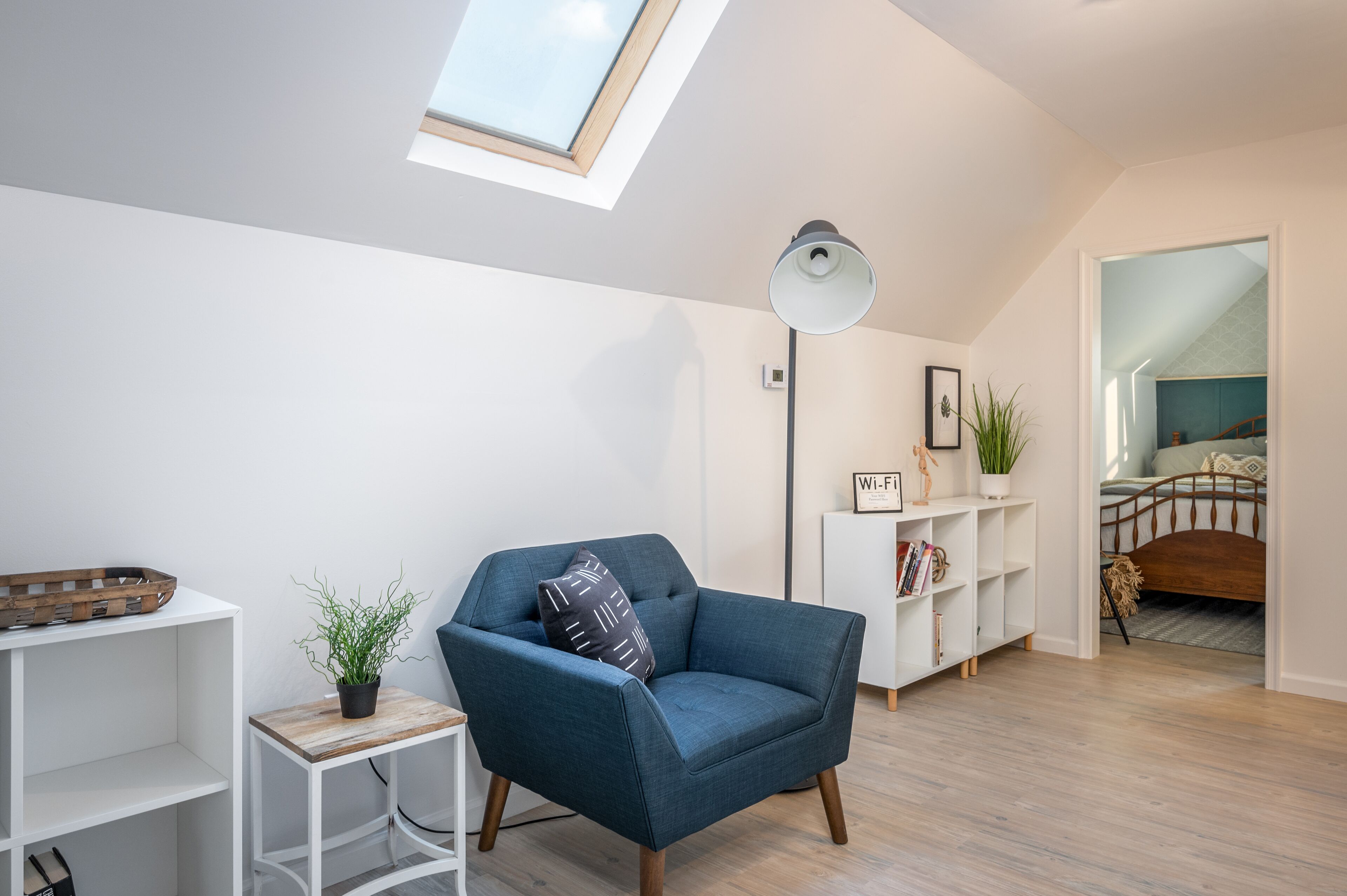 Quarto moderno com claraboia, uma poltrona azul, mesa de cabeceira com planta e cama de madeira.