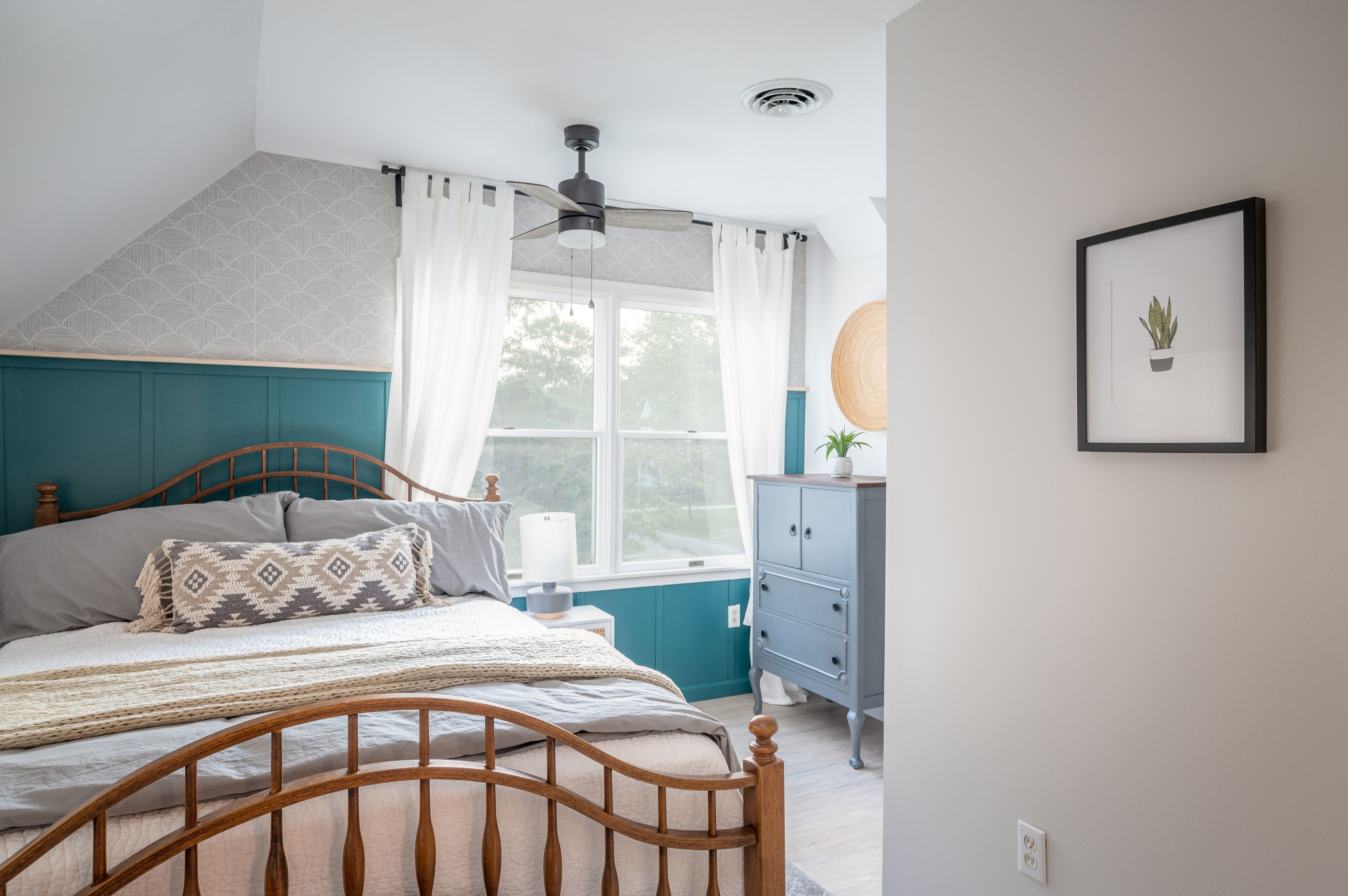 A bedroom with a wooden bed, a window with curtains, a ceiling fan, a dresser, and a framed picture on the wall.