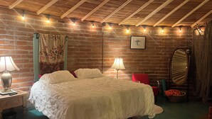 A cozy room with a brick wall, wooden ceiling beams, a large bed with white bedding, two bedside lamps, a red chair, a mirror, and a framed picture on the wall.
