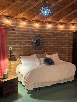 A cozy bedroom with a brick wall, a star-shaped hanging light, a round mirror, a wooden nightstand with a lamp, and a bed with white bedding and pillows. - Elegant magical rustic round house by Saguaro National Park  (Tucson)