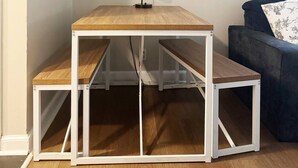 Uma pequena mesa de jantar de madeira com estrutura branca e bancos, colocada num canto de um quarto com um sofá azul e um quadro na parede.