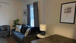 Uma sala de estar com sofá, mesa, abajur, cadeira e uma janela com cortinas.