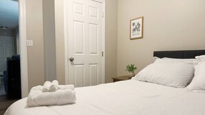 Cama bem arrumada com lençóis brancos, mesa de cabeceira com uma planta e porta com um quadro na parede.
