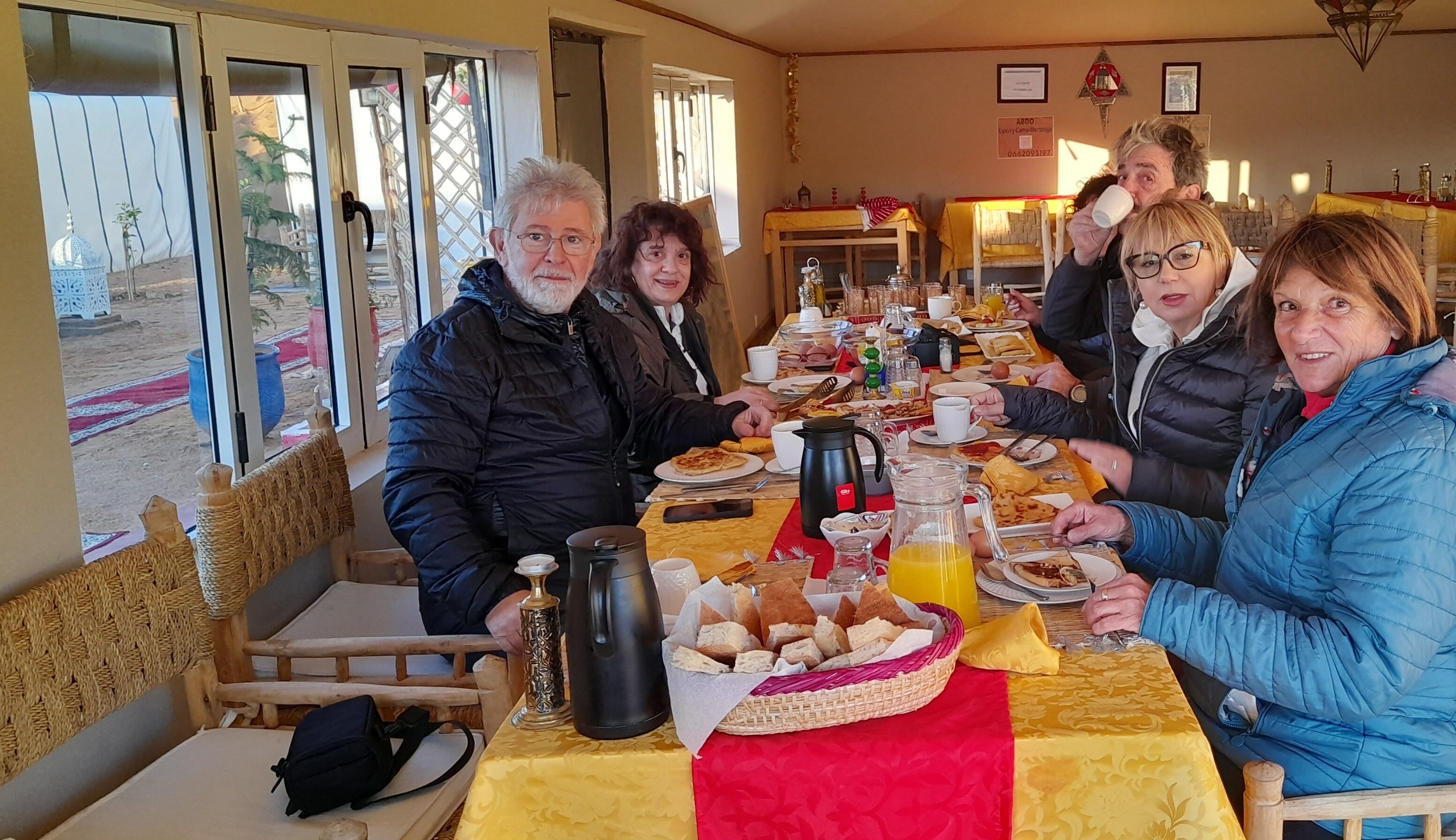 Un groupe de personnes est assis à une table, devant un assortiment de produits pour le petit-déjeuner, une cafetière et un panier de pain.