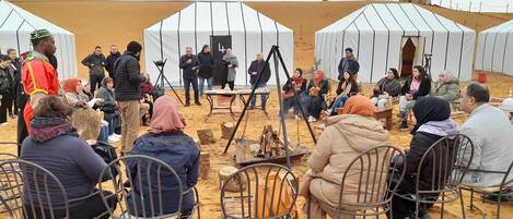 A group of people seated in a semi-circular arrangement around a central fire pit, with tents in the background.