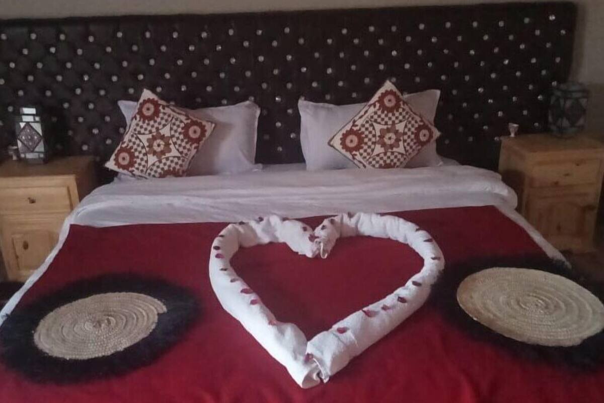 A neatly made bed with a heart-shaped towel and two circular woven placemats on a red bedspread.