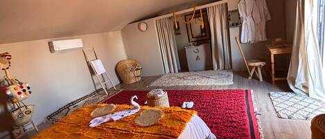 A cozy room with a large bed, a small table, and a chair. There is a window with curtains, a wall-mounted air conditioner, and a star-shaped hanging light.