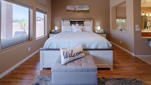 A bedroom with a large bed, a bench with a 'Welcome' pillow, a painting on the wall, and a bathroom visible through an archway. - Rare 3 FULL bathroom home in golf community W/ private pool. Experienced Host. (Mesa)