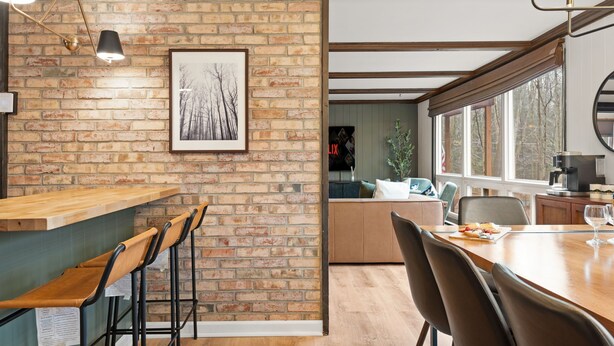 Una cocina moderna con pared de ladrillo, taburetes de bar de madera, una mesa de comedor con sillas y un ventanal que proporciona luz natural.