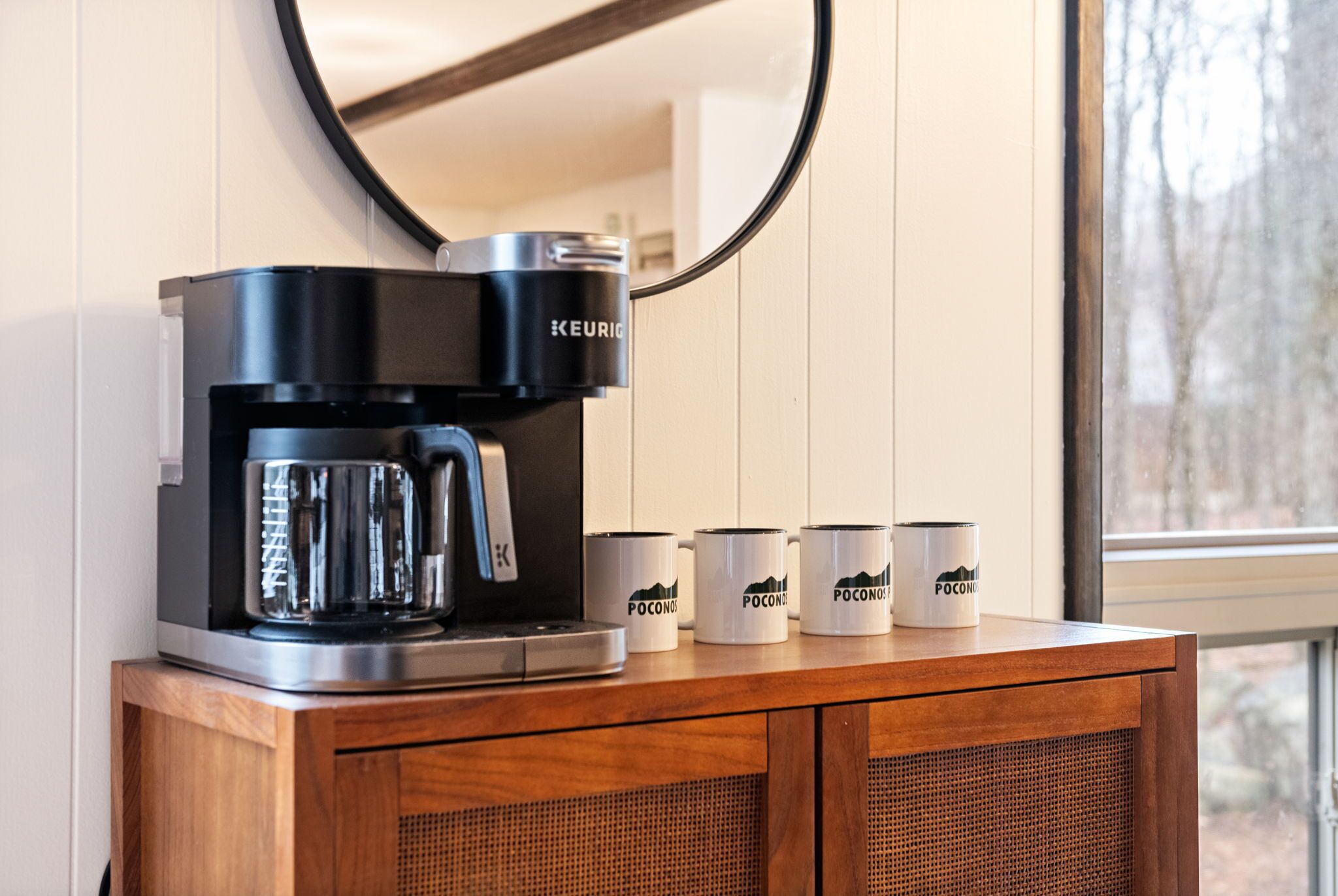 A Keurig coffee maker on a wooden cabinet with four white mugs labeled "POCONOS" and a mirror above it.