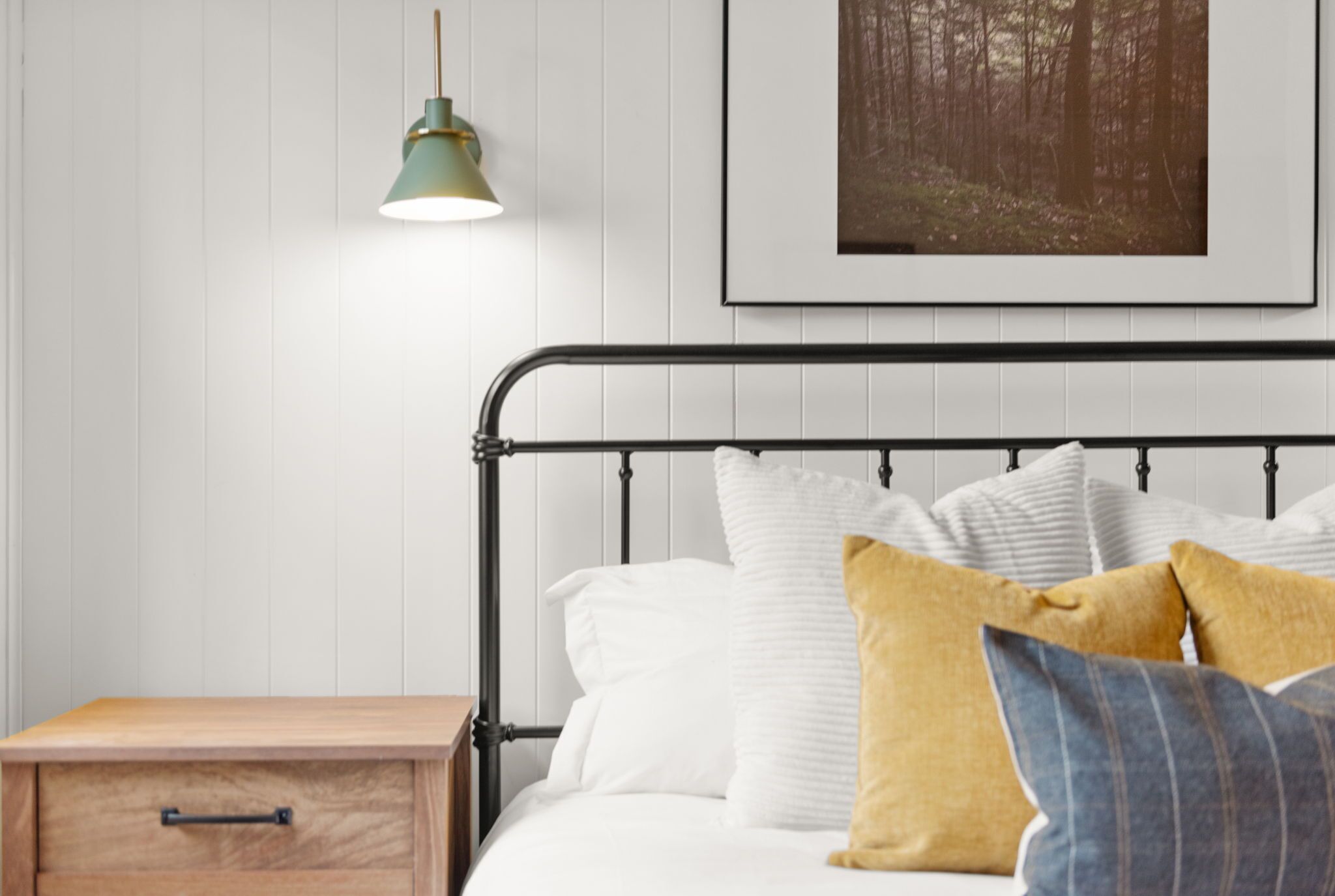 Una cama bien hecha con almohadillas blancas y mostaza, una lámpara verde montada en la pared y una imagen enmarcada de un bosque encima.