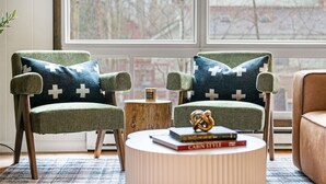 Dos sillones con cojines de estampado cruce, una mesita de madera y una mesa de centro redonda con libros y un objeto decorativo.