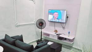 A modern living room with a dark sofa, a small coffee table, a TV on the wall, a fan, and a potted plant. - Motalvin Court  (Lagos)