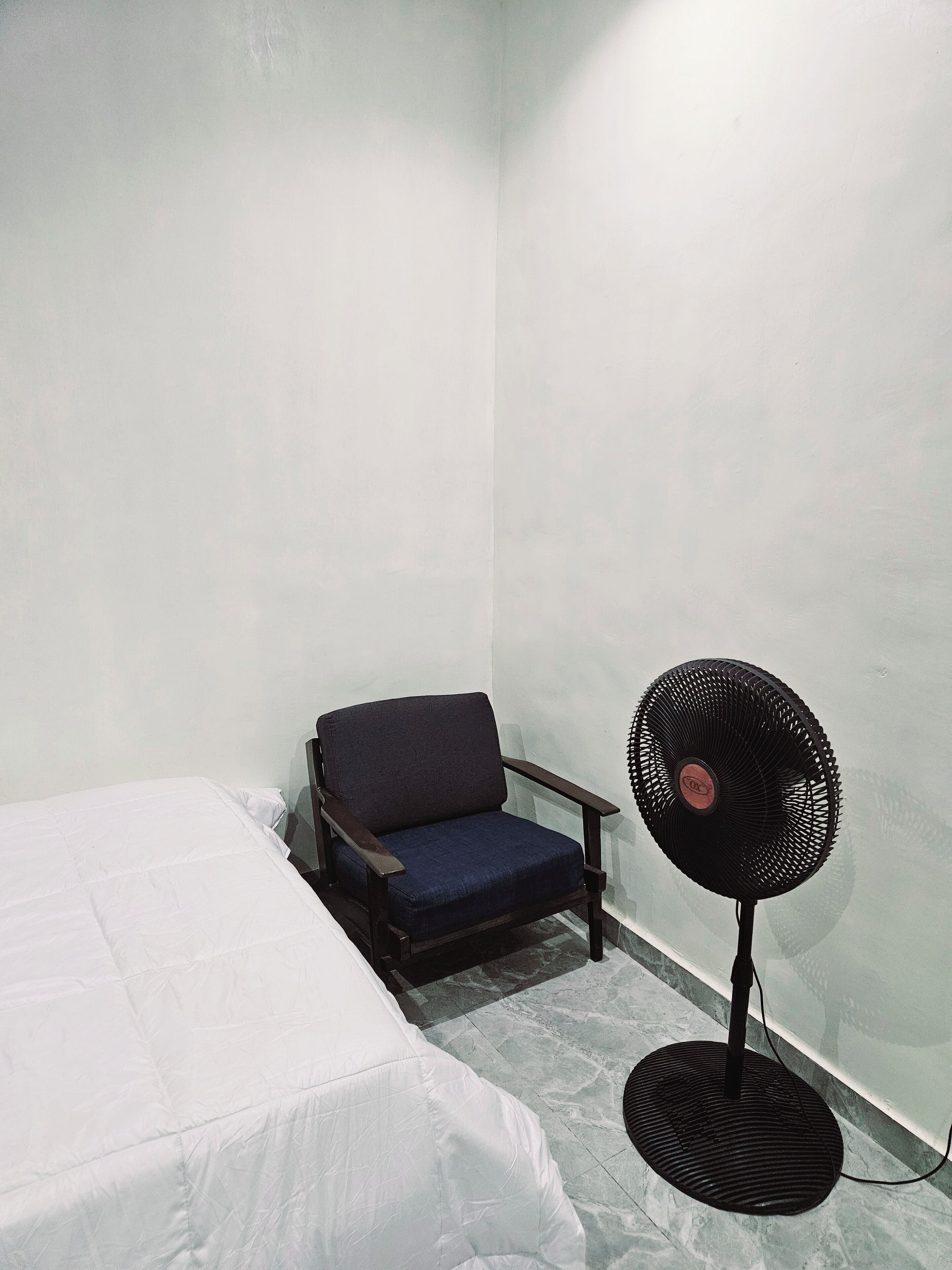 A small room with a bed, a chair, and a standing fan.