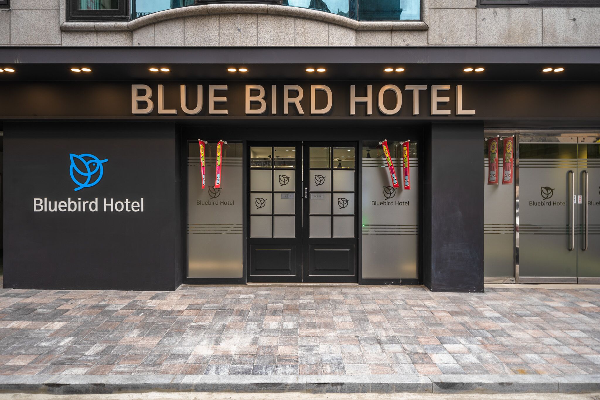 L’extérieur de l’hôtel Bluebird se distingue par sa façade moderne, ses grandes portes vitrées et son logo bien visible.
