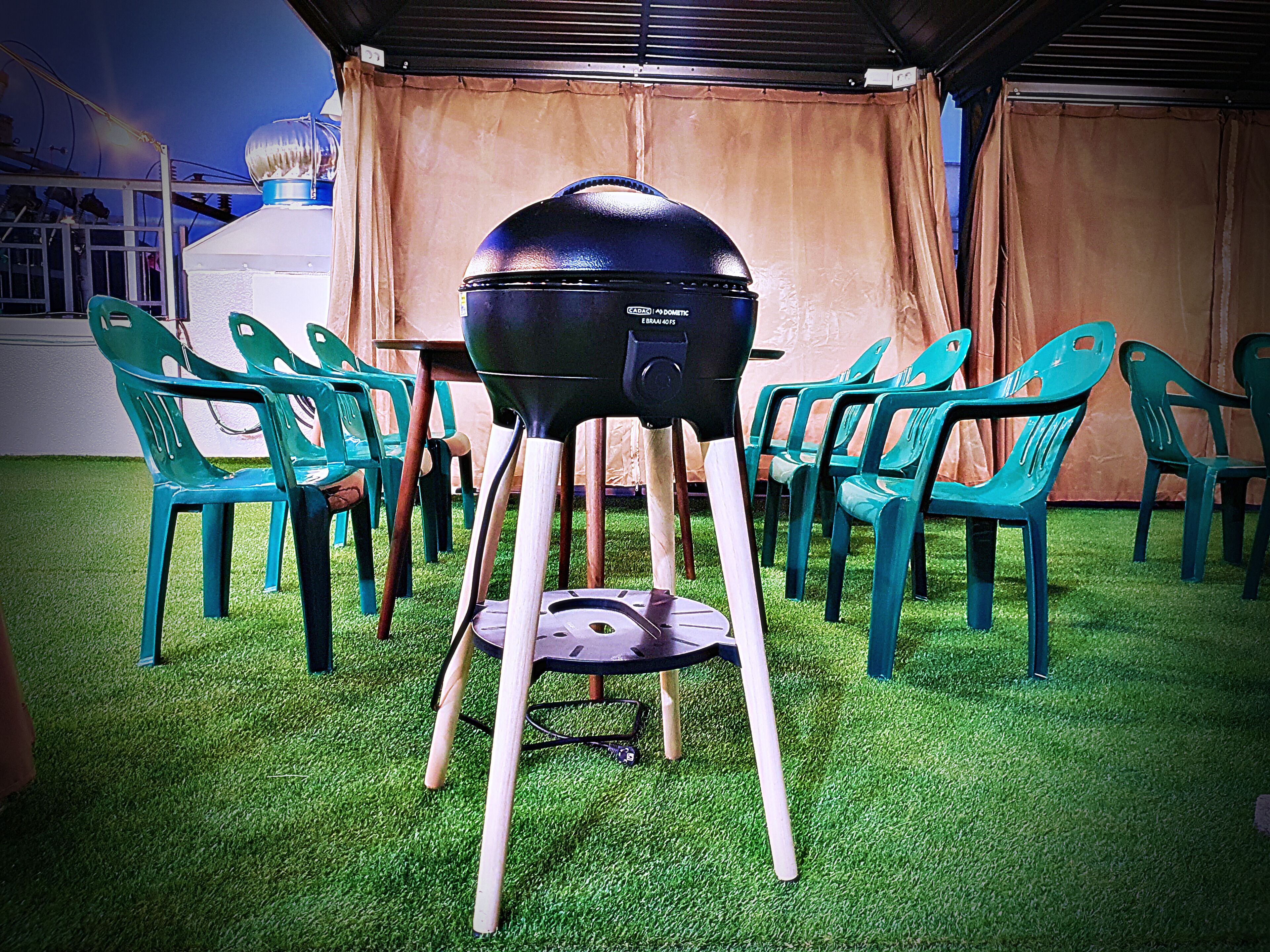 Un barbecue doté d’un couvercle en forme de dôme, entouré de chaises en plastique vertes posées sur du gazon synthétique.