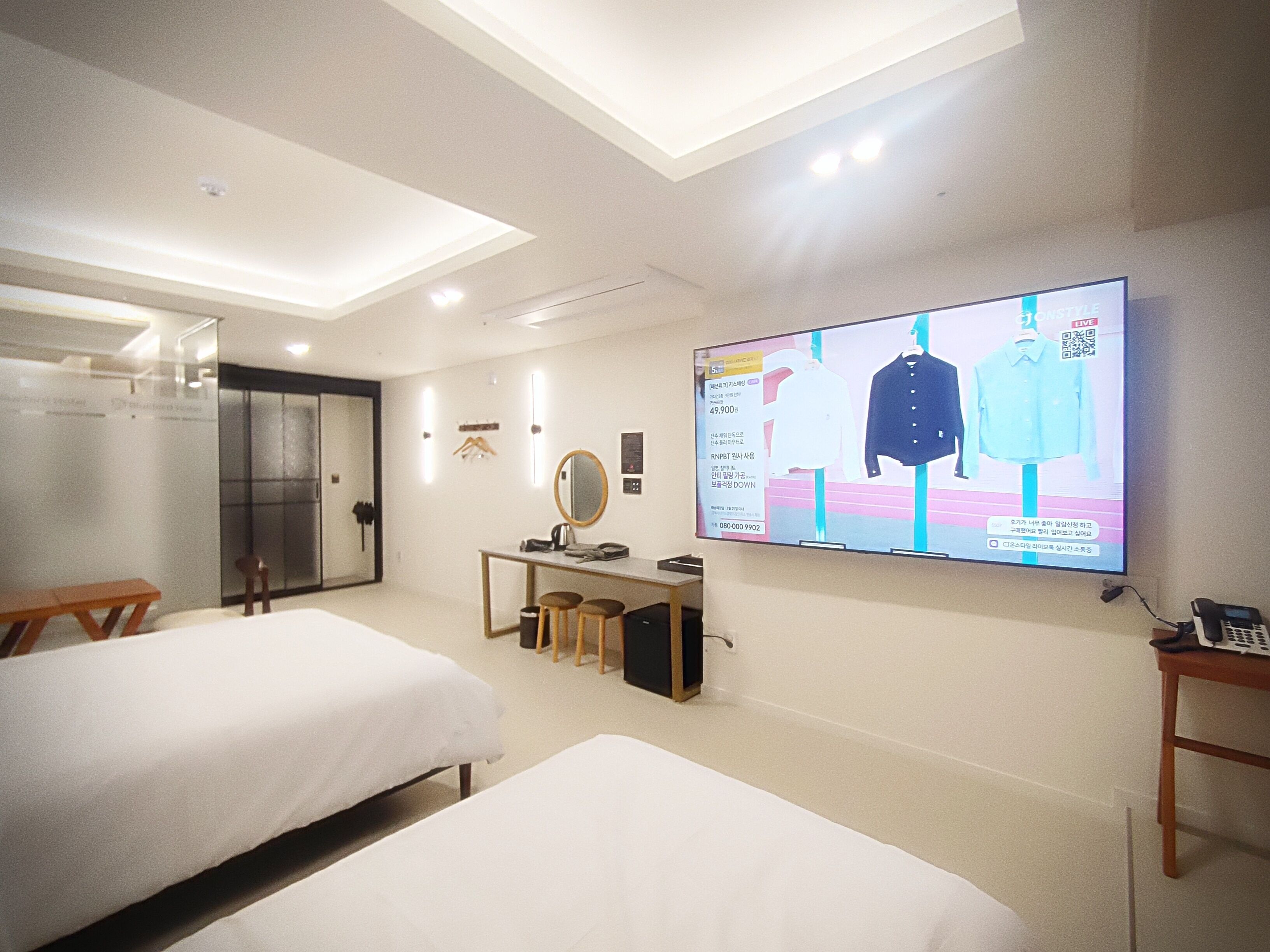 A hotel room with two beds, a desk with a computer, a TV displaying a clothing advertisement, and a mirror on the wall.