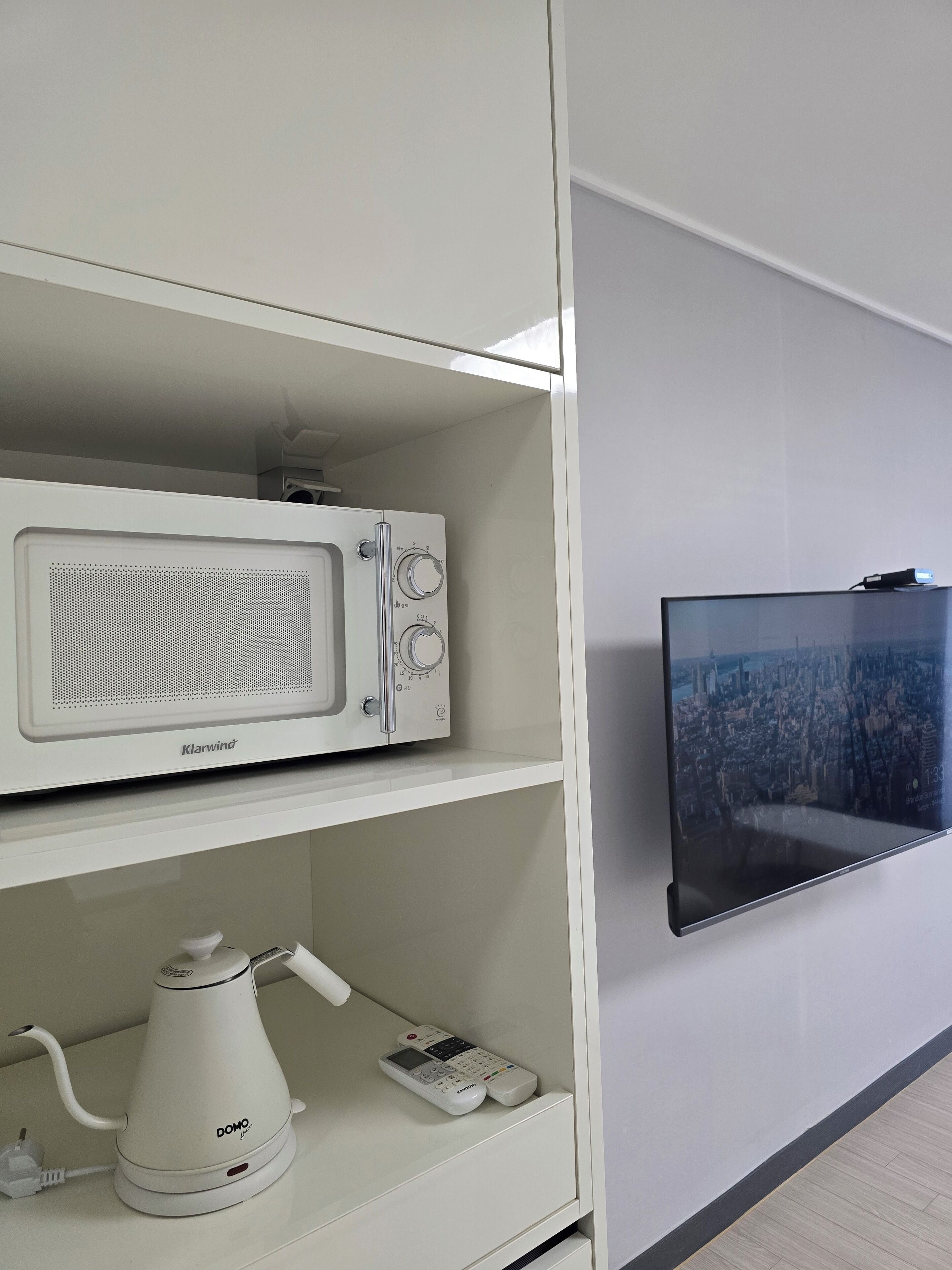 Un meuble blanc contenant un four à micro-ondes, une machine à café et une télécommande sur l’étagère inférieure. Un téléviseur à écran plat est fixé au mur, à droite.