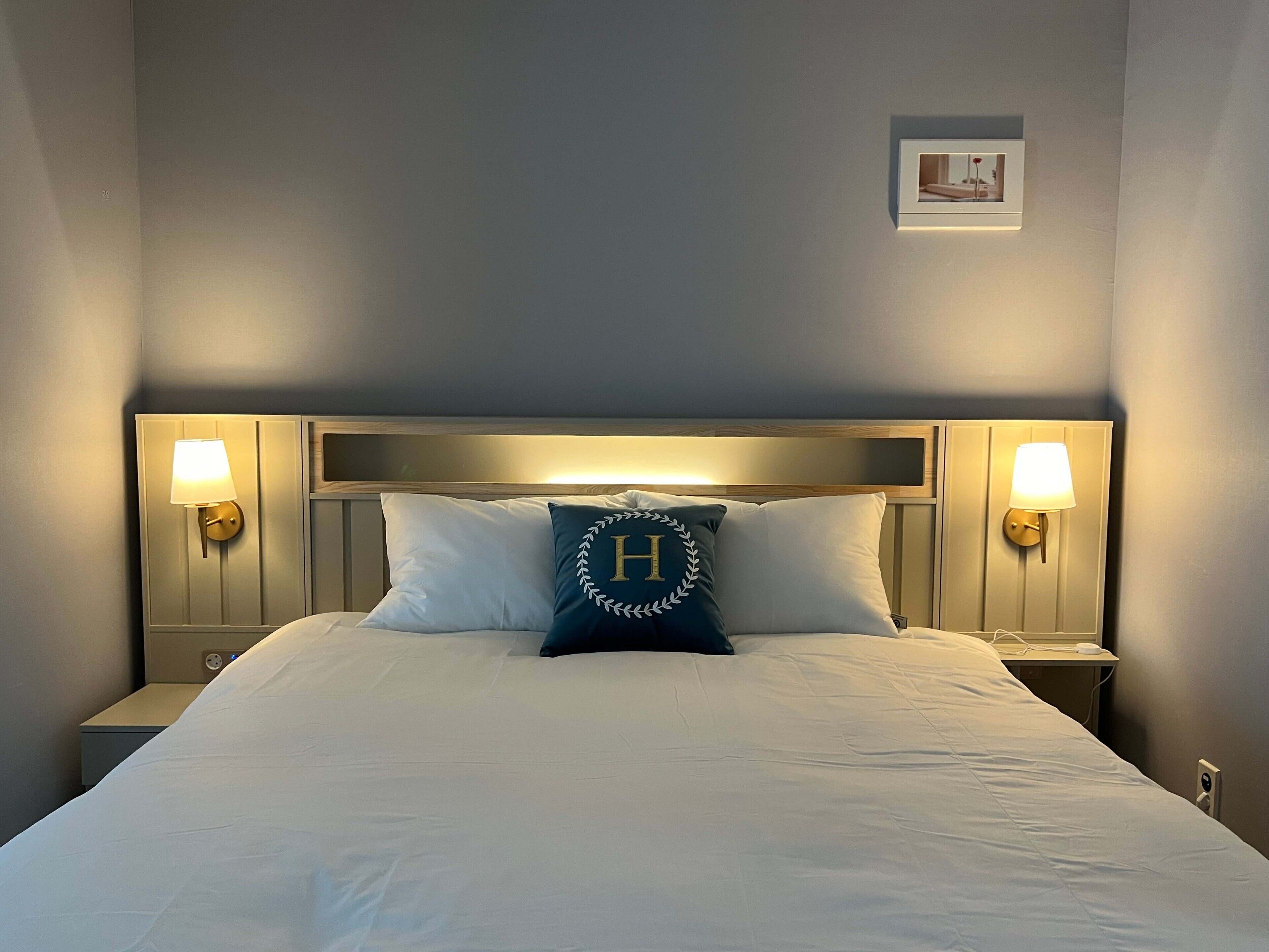 A neatly made bed with white linens, a pillow with a monogram, and two wall-mounted lamps on either side.