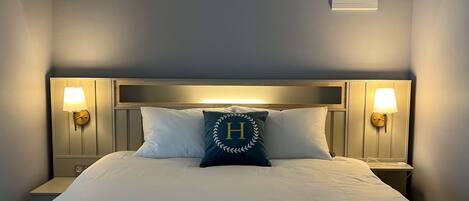 A neatly made bed with white linens, a pillow with a monogram, and two wall-mounted lamps on either side.