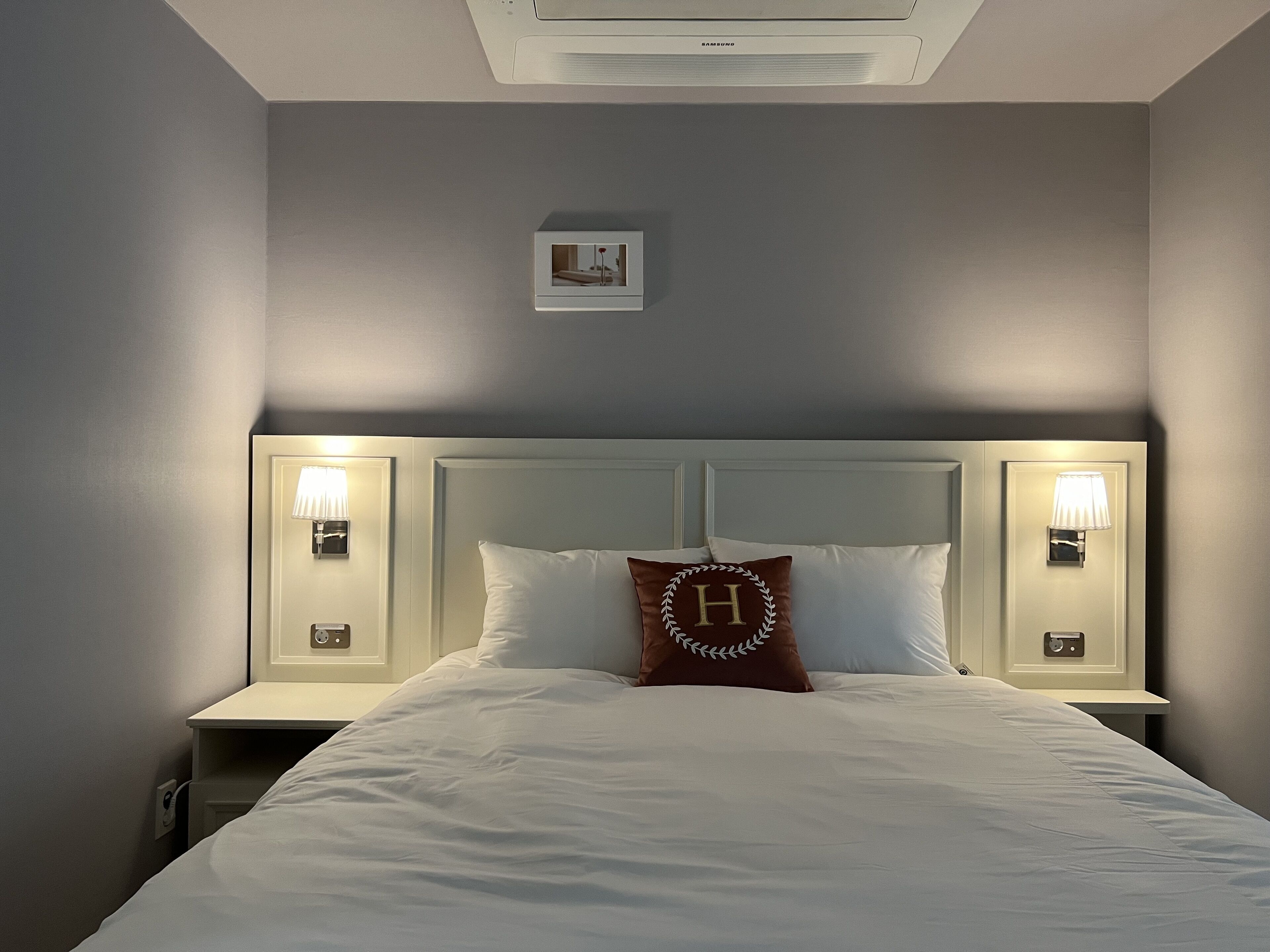 A neatly made bed with white bedding, a brown pillow with a monogram, and two bedside lamps on either side.